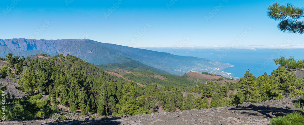 Panoramatic view of volcanic landscape with lush green pine trees, colorful volcanoes and white clouds at path Ruta de los Volcanes, hiking trail. La Palma, Canary Islands, Spain, Blue sky background