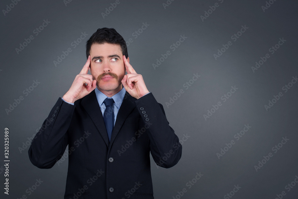 Dreamy Caucasian male with thoughtful expression, looks away, keeps hand near face, thinks about something pleasant, poses against gray wall. Dreamy Caucasian male with thoughtful expression, looks away, keeps hand near face, thinks about something pleasant, poses against gray wall.