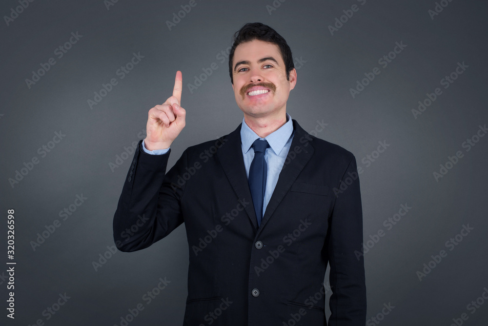 Young caucasian businessman standing against gray wall showing and pointing up with fingers number one while smiling confident and happy. Young caucasian businessman standing against gray wall showing and pointing up with fingers number one while smiling confident and happy.