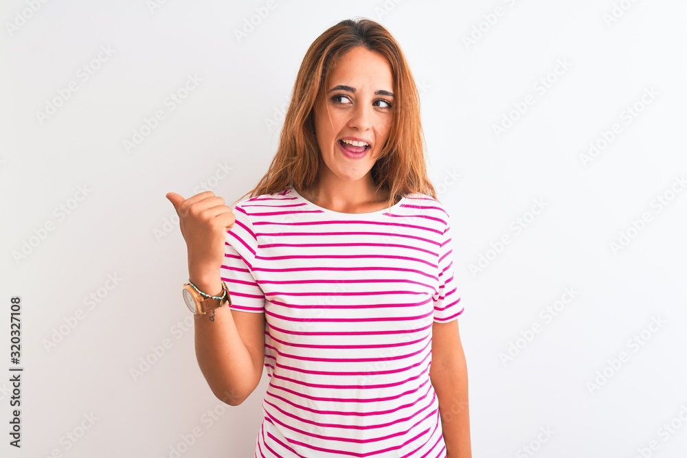 Young redhead woman wearing striped casual t-shirt stading over white isolated background smiling with happy face looking and pointing to the side with thumb up. Young redhead woman wearing striped casual t-shirt stading over white isolated background smiling with happy face looking and pointing to the side with thumb up.