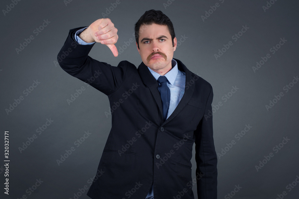 Discontent European man shows disapproval sign, keeps thumb down, expresses dislike, frowns face in discontent, dressed in white shirt, isolated over gray background. Body language concept. Discontent European man shows disapproval sign, keeps thumb down, expresses dislike, frowns face in discontent, dressed in white shirt, isolated over gray background. Body language concept.