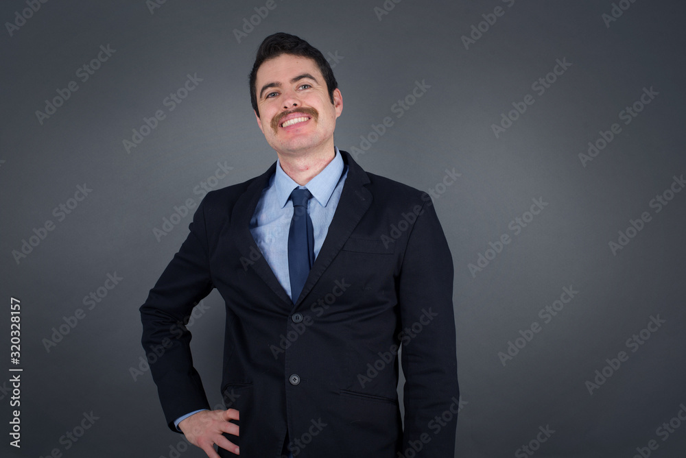 Studio shot of cheerful businessman keeps hand on hip, smiles broadly, anticipates for surprise prepared by friends on his birthday, isolated over gray background. Studio shot of cheerful businessman keeps hand on hip, smiles broadly, anticipates for surprise prepared by friends on his birthday, isolated over gray background.