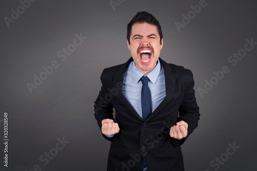 Close up portrait of stylish handsome caucasian businessman raising fists up isolated on shine grey background big eyes and wide open mouth. © Jihan