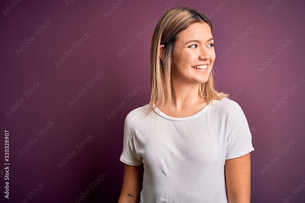 Fototapeta premium Young beautiful blonde woman wearing casual white t-shirt over purple isolated background looking away to side with smile on face, natural expression. Laughing confident.