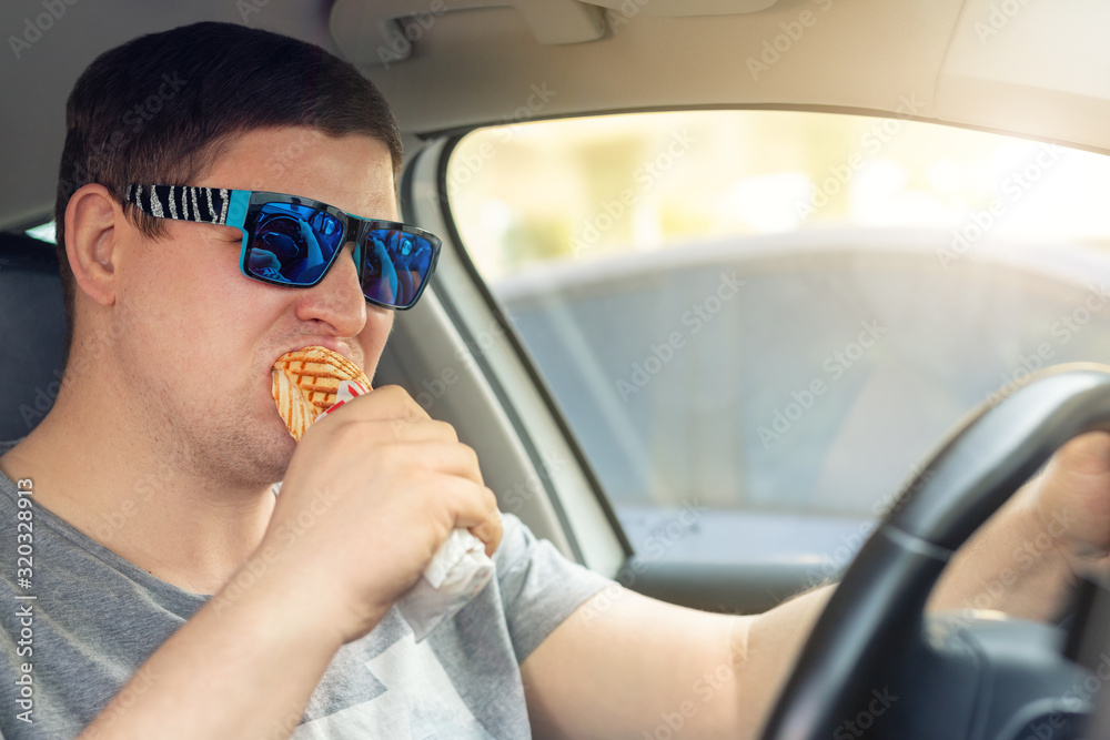 Adult male caucasian person eating hot dog fast food snack while driving car on sunny day. Men hsving food while driving steering wheel. Distracted man in vehicle. Bad unhealthy habit. Junk food go