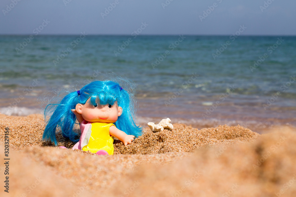 Obraz premium girl doll with blue hair on the sand on the beach with a piece of coral Playing in the sand on the beach by the sea in summer during travel or vacation A doll with blue hair is sitting on the sand