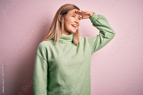 Young beautiful blonde woman wearing winter wool sweater over pink isolated background very happy and smiling looking far away with hand over head. Searching concept. © Krakenimages.com