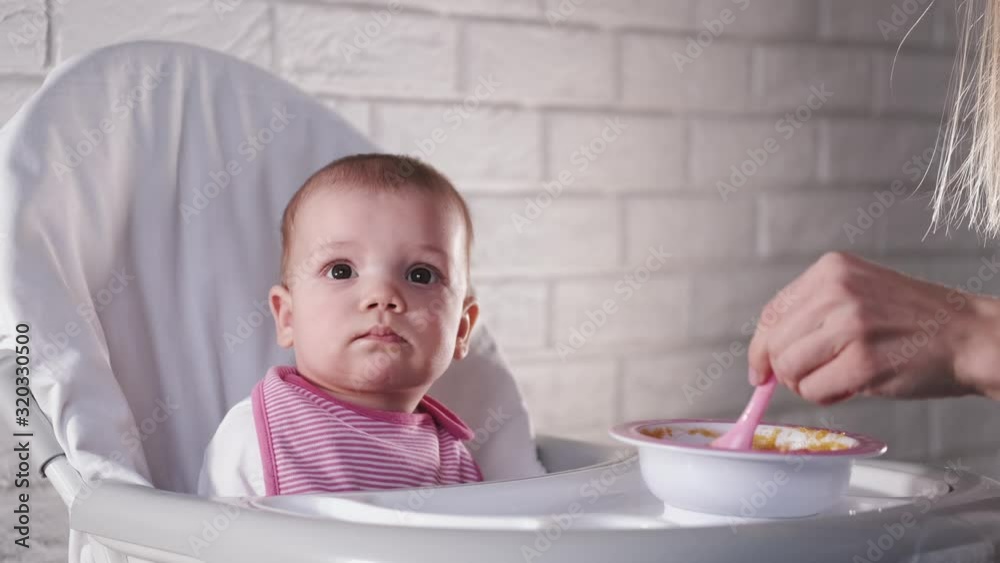 A baby in a bib is being fed by it’s mother with a spoon. The newborn is having a meal in a chair for feeding with food remains on it’s mouth.