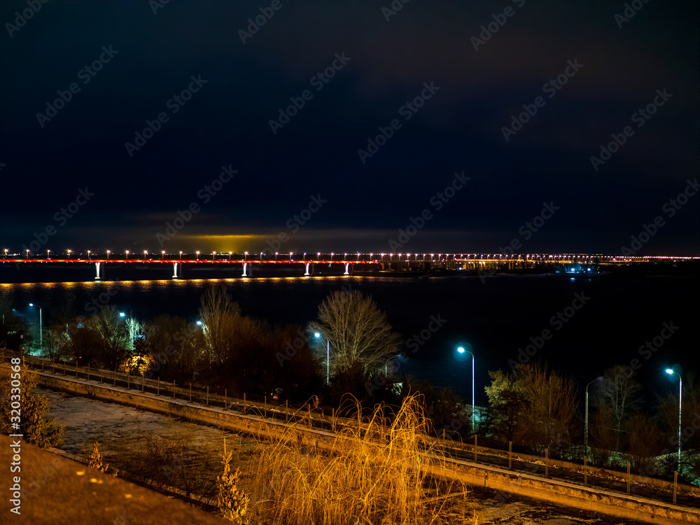 long automobile bridge over large river illuminated by multi-colored lights. Cars driving along embankment of river. little traffic late in evening. Trees in foreground