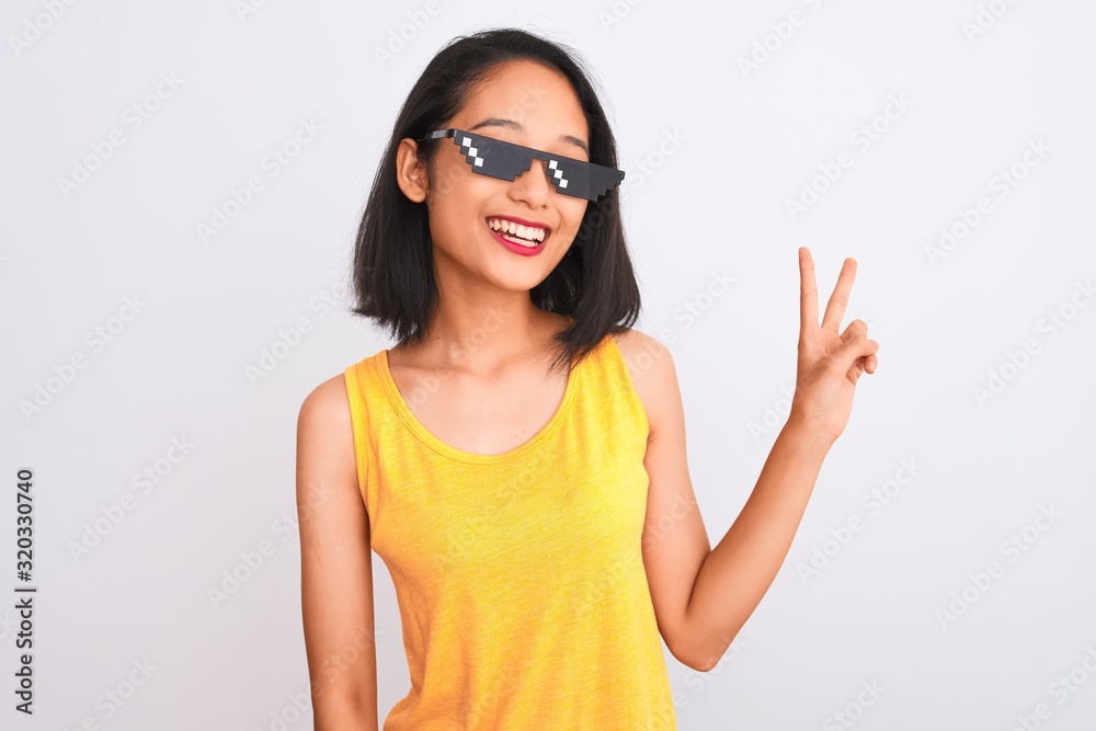 Young beautiful chinese woman wearing thug life sunglasses over isolated white background smiling with happy face winking at the camera doing victory sign. Number two. Young beautiful chinese woman wearing thug life sunglasses over isolated white background smiling with happy face winking at the camera doing victory sign. Number two.