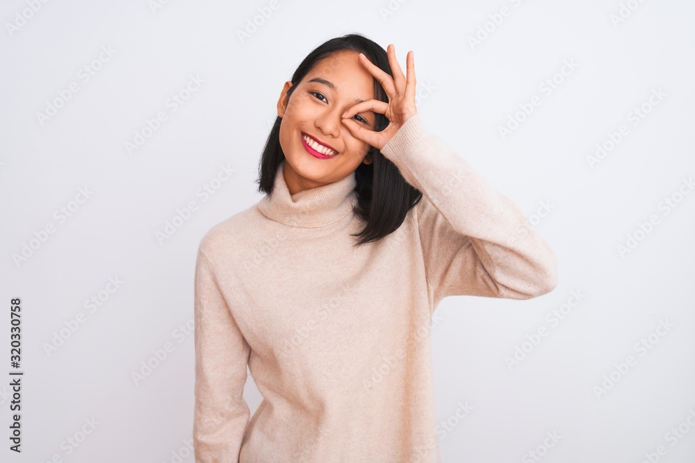 Fototapeta premium Young chinese woman wearing turtleneck sweater standing over isolated white background doing ok gesture with hand smiling, eye looking through fingers with happy face.