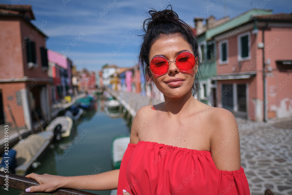  Young fashionable woman in romantic red  dress  with braid hairstyle, posing near colorful houses in Burano Island, Venice, Italy