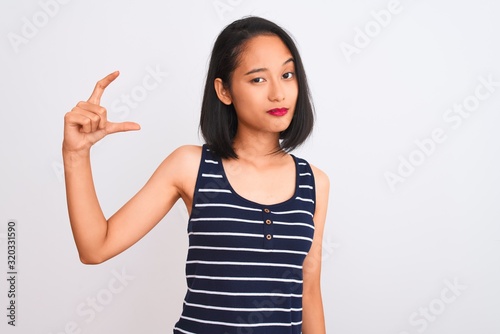 Young chinese woman wearing striped t-shirt standing over isolated white background smiling and confident gesturing with hand doing small size sign with fingers looking and the camera. Measure