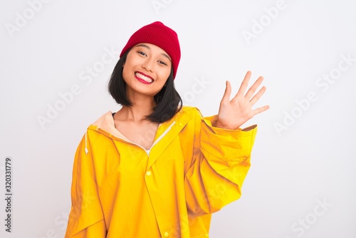 Young beautiful chinese woman wearing raincoat and wool cap over isolated white background showing and pointing up with fingers number five while smiling confident and happy. © Krakenimages.com