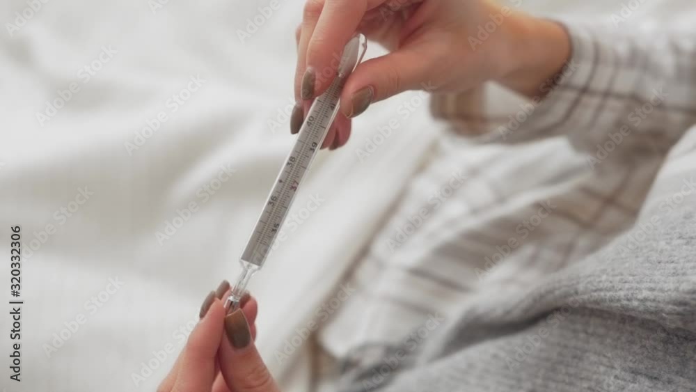 Close-up scene of a girl who is lying in the bed and holding a mercury thermometer in her hand. She has a fever.