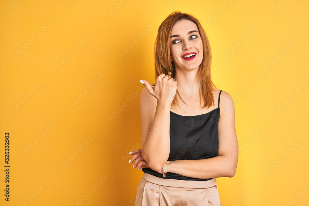 Redhead caucasian business woman standing over yellow isolated background smiling with happy face looking and pointing to the side with thumb up. Redhead caucasian business woman standing over yellow isolated background smiling with happy face looking and pointing to the side with thumb up.