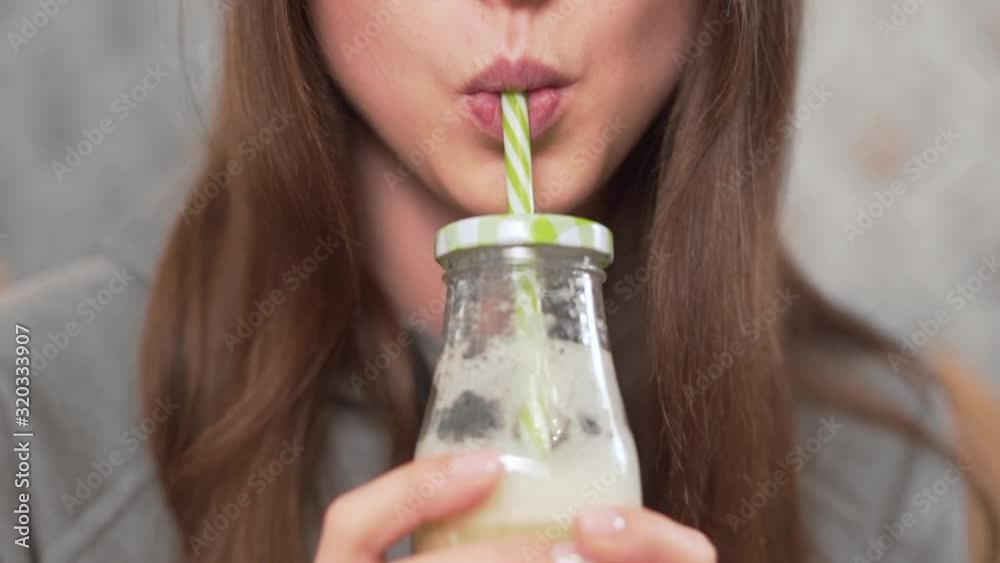 Close-up scene of a beautiful girl who is sitting at home on the couch, drinking a homemade milkshake from a bottle with a straw.