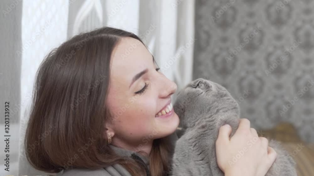 Beautiful girl is sitting in her room, holding a grey cat on her hands and smiling. Cat is licking her face. Beautiful girl is sitting in her room, holding a grey cat on her hands and smiling. Cat is licking her face.