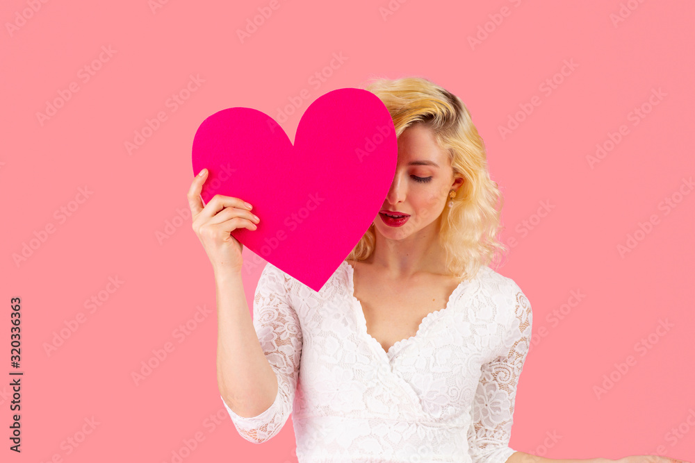 Studio portrait of a beautiful young woman holding pink heart covering one eye and looking down, love and dating concept Studio portrait of a beautiful young woman holding pink heart covering one eye and looking down, love and dating concept