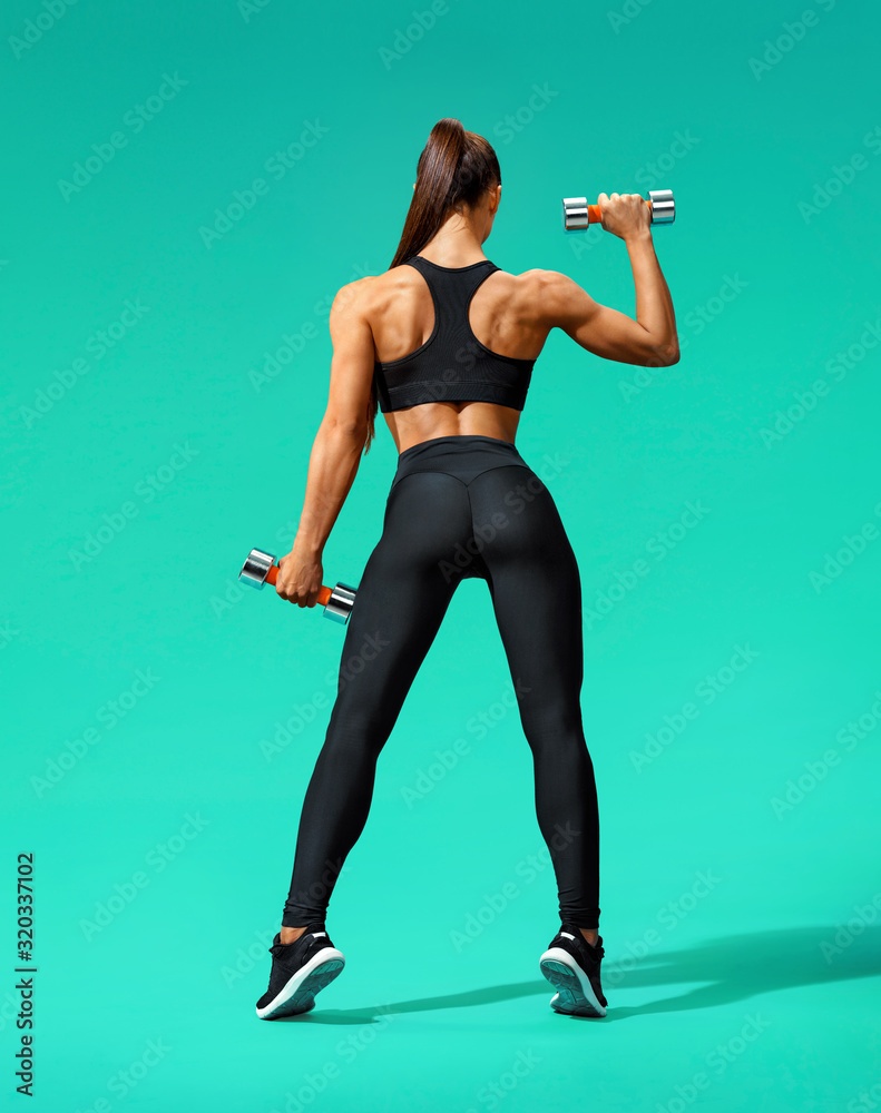 Strong woman working out with dumbbells, flexing her arm. Photo of sporty woman in black sportswear on turquoise background. Rear view. Full length Strong woman working out with dumbbells, flexing her arm. Photo of sporty woman in black sportswear on turquoise background. Rear view. Full length