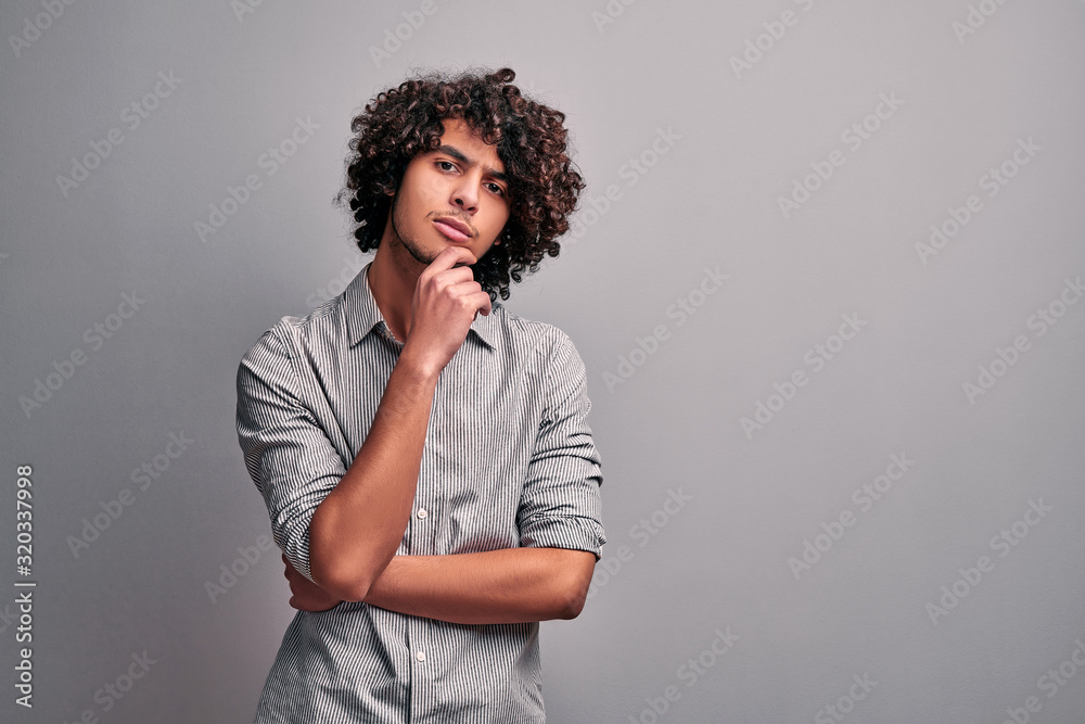 Handsome eastern mindful student holding hand on his chin while thinking, standing against gray wall. Handsome eastern mindful student holding hand on his chin while thinking, standing against gray wall.