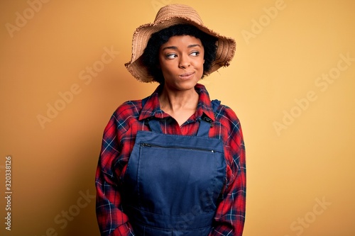 Young African American afro farmer woman with curly hair wearing apron and hat smiling looking to the side and staring away thinking.