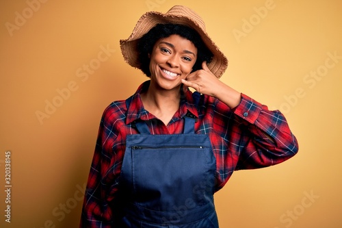 Young African American afro farmer woman with curly hair wearing apron and hat smiling doing phone gesture with hand and fingers like talking on the telephone. Communicating concepts.