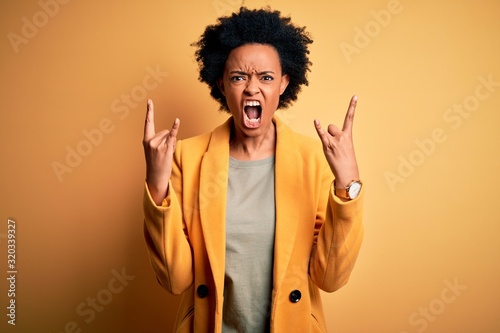 Young beautiful African American afro businesswoman with curly hair wearing yellow jacket shouting with crazy expression doing rock symbol with hands up. Music star. Heavy concept.