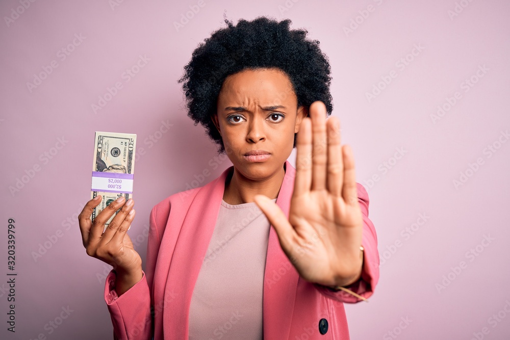 Young African American afro businesswoman with curly hair holding dollars banknotes with open hand doing stop sign with serious and confident expression, defense gesture Young African American afro businesswoman with curly hair holding dollars banknotes with open hand doing stop sign with serious and confident expression, defense gesture