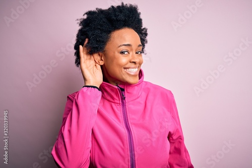 Young African American afro sportswoman with curly hair wearing sportswear doin sport smiling with hand over ear listening an hearing to rumor or gossip. Deafness concept.