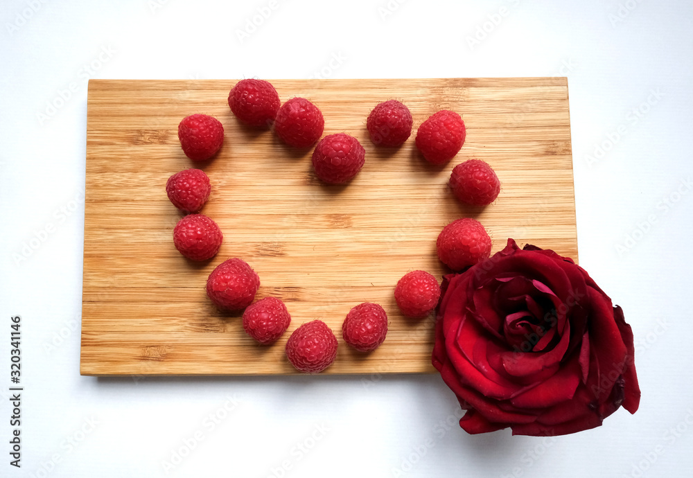Fototapeta premium Tasty juicy ripe fresh red raspberry berries in the heart shaped form on a wooden cutting board and a red rose flowe