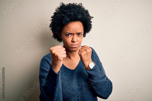 Young beautiful African American afro woman with curly hair wearing casual sweater Ready to fight with fist defense gesture, angry and upset face, afraid of problem