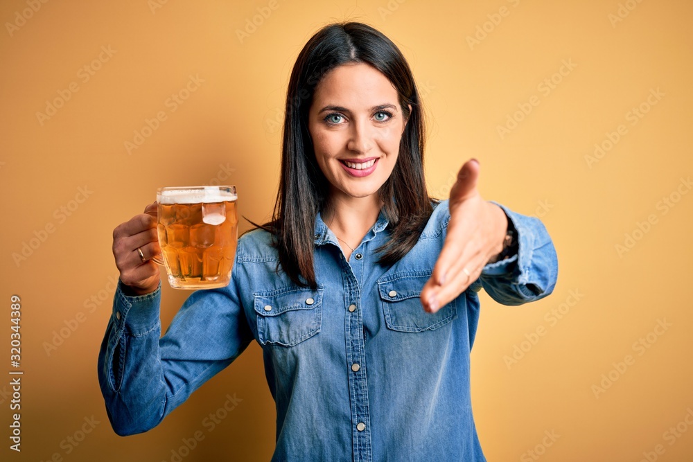 Young woman with blue eyes drinking jar of beer standing over isolated yellow background smiling friendly offering handshake as greeting and welcoming. Successful business. Young woman with blue eyes drinking jar of beer standing over isolated yellow background smiling friendly offering handshake as greeting and welcoming. Successful business.