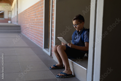 Schoolboy sitting on a step by a wall in the schoolyard at elementary school using a tablet computer