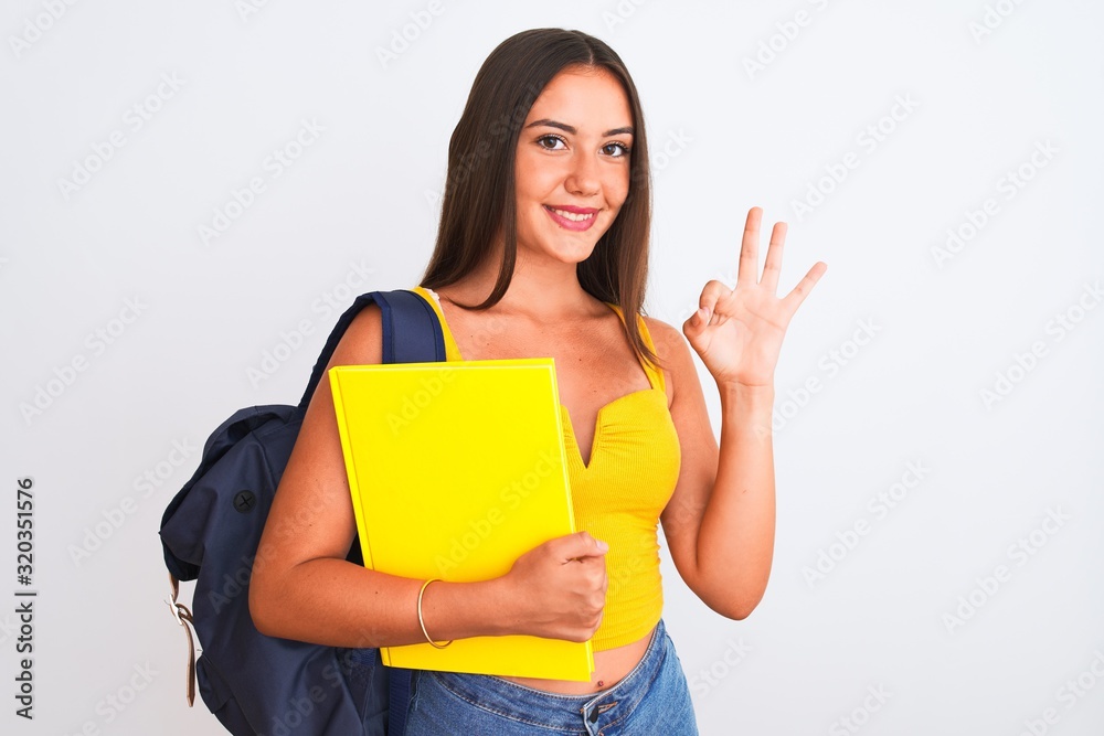 Beautiful student girl wearing backpack holding notebook over isolated white background doing ok sign with fingers, excellent symbol Beautiful student girl wearing backpack holding notebook over isolated white background doing ok sign with fingers, excellent symbol