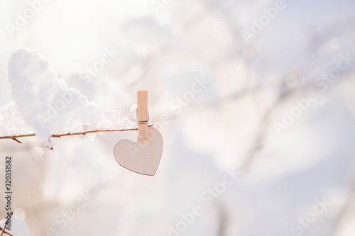 wooden heart hanging on a clothespin on a snow branch, glare . The concept of Valentine's Day .