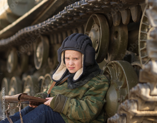 a boy sits near a tank on the street in the form of a tankman of a military green color with a tablet in his hands photo