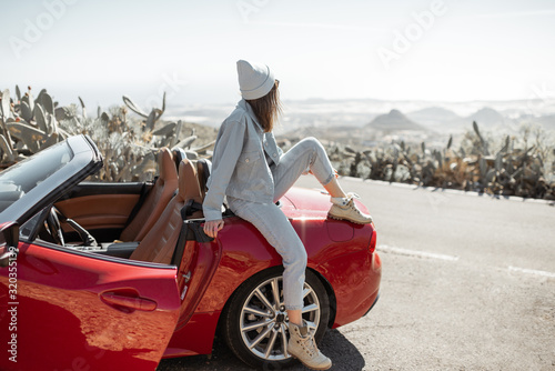 Lifestyle portrait of a stylish woman dressed casually in jeans and hat sitting on the car, enjoying road trip on the island