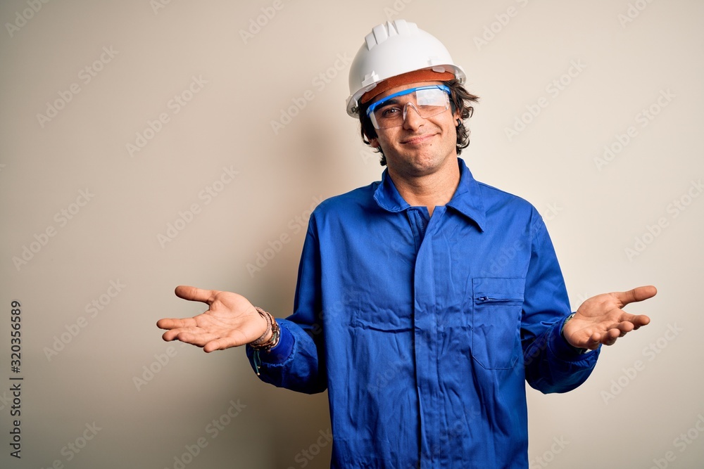 Young constructor man wearing uniform and security helmet over isolated white background clueless and confused expression with arms and hands raised. Doubt concept.