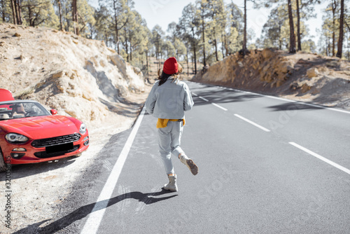 Lifestyle portrait of a carefree woman running on the beautiful mountain road, having a stop while traveling by car on the island. Happy road trip concept