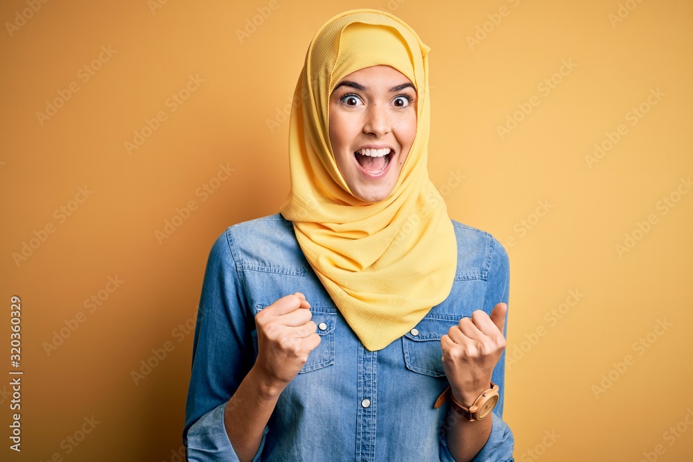 Young beautiful girl wearing muslim hijab standing over isolated yellow background celebrating surprised and amazed for success with arms raised and open eyes. Winner concept. Young beautiful girl wearing muslim hijab standing over isolated yellow background celebrating surprised and amazed for success with arms raised and open eyes. Winner concept.