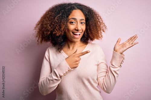 Young african american woman with afro hair wearing casual sweater over pink background amazed and smiling to the camera while presenting with hand and pointing with finger.