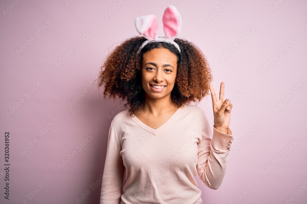 Young african american woman with afro hair wearing bunny ears over pink background smiling looking to the camera showing fingers doing victory sign. Number two.