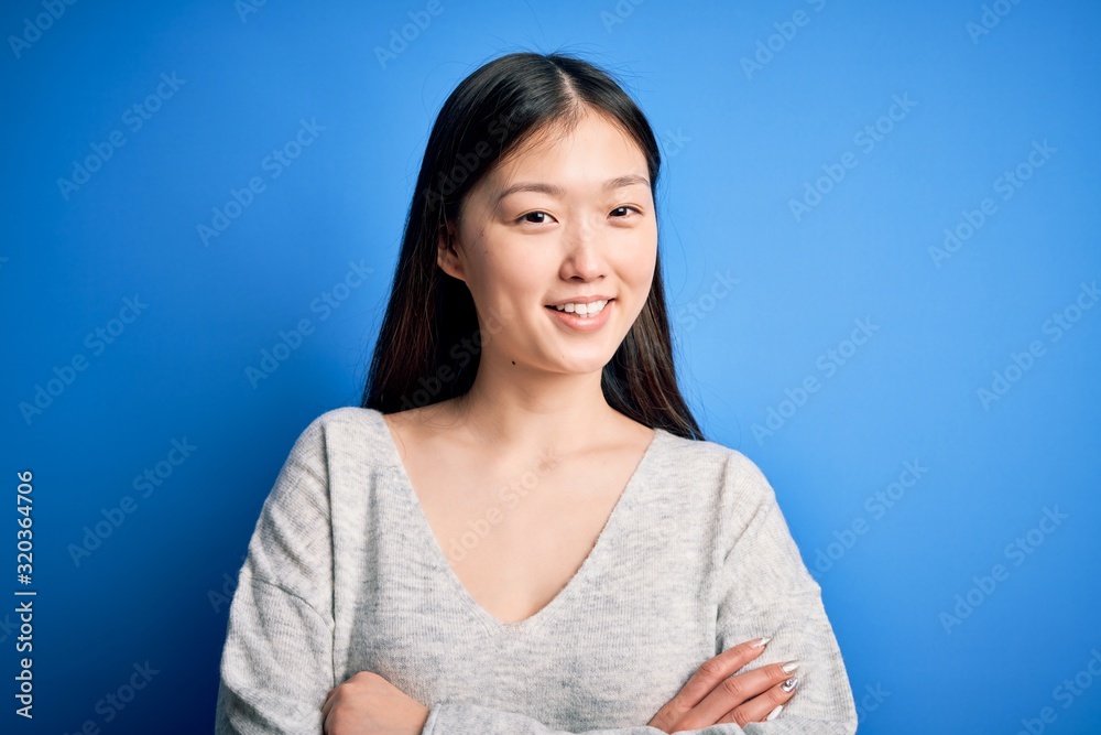 Young beautiful asian woman wearing casual sweater standing over blue isolated background happy face smiling with crossed arms looking at the camera. Positive person. Young beautiful asian woman wearing casual sweater standing over blue isolated background happy face smiling with crossed arms looking at the camera. Positive person.
