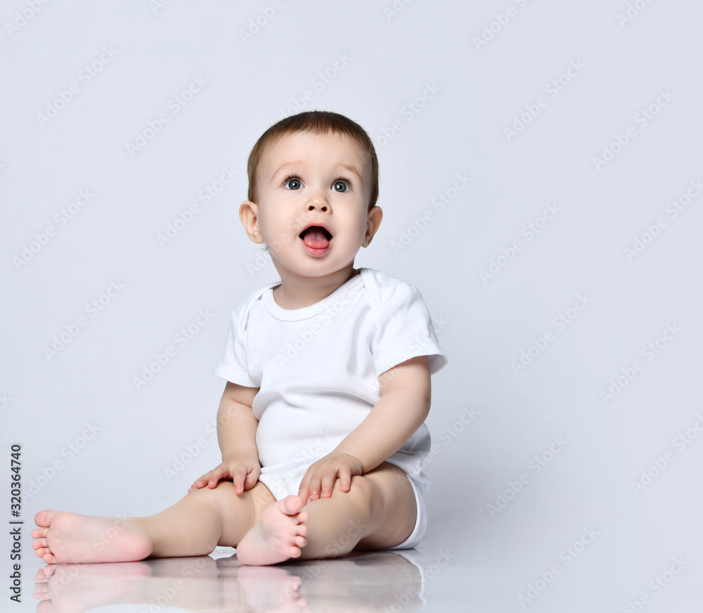Happy baby boy toddler in white bodysuit is sitting on the floor with open mouth looking up on gray with copy space Happy baby boy toddler in white bodysuit is sitting on the floor with open mouth looking up on gray with copy space