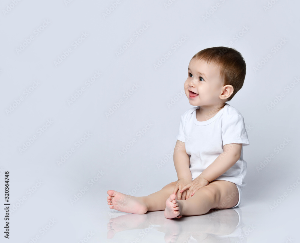 Happy smiling baby boy toddler in white bodysuit is sitting sideways on the floor looking up at copy space on a gray