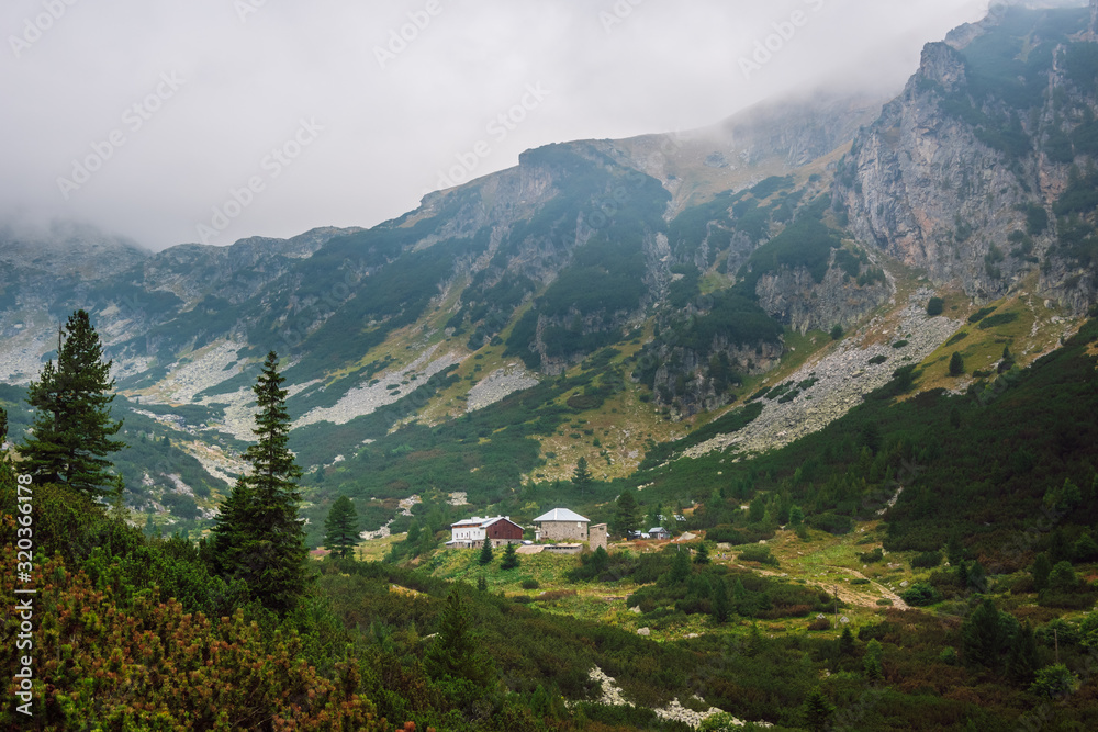 View of a green mountain valley with а refuge, steep rocky slopes, covered with coniferous shrubs and descending fog to the valley, refuge Malyovitsa, Rila mountain, Bulgaria.