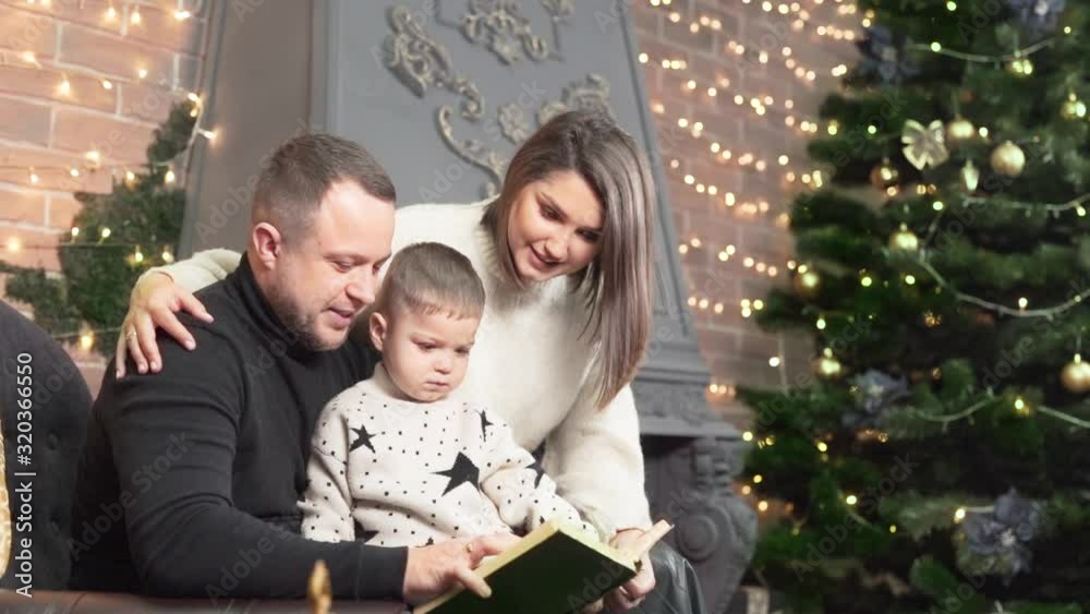 Cute family is preparing a christmas tree in their home before New Year. Young mother, father and their little son are sitting in a chair and reading a book. Cute family is preparing a christmas tree in their home before New Year. Young mother, father and their little son are sitting in a chair and reading a book.