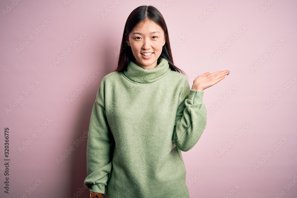 Young beautiful asian woman wearing green winter sweater over pink solated background smiling cheerful presenting and pointing with palm of hand looking at the camera. Young beautiful asian woman wearing green winter sweater over pink solated background smiling cheerful presenting and pointing with palm of hand looking at the camera.