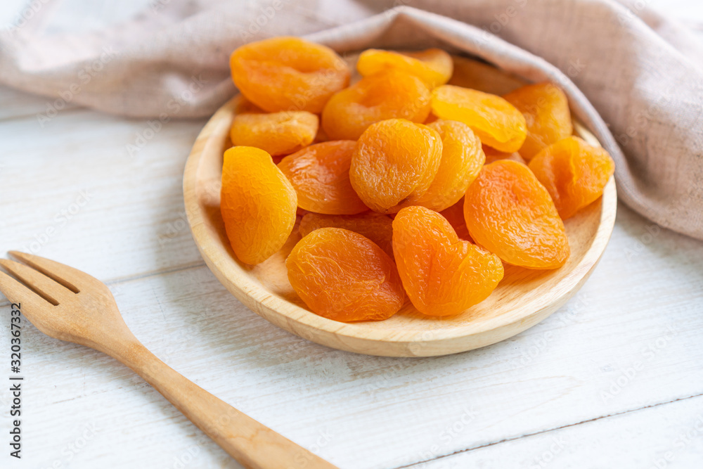 Dried apricots in wooden plate on white wooden background with table cloth and fork, close up. Dried apricots in wooden plate on white wooden background with table cloth and fork, close up.